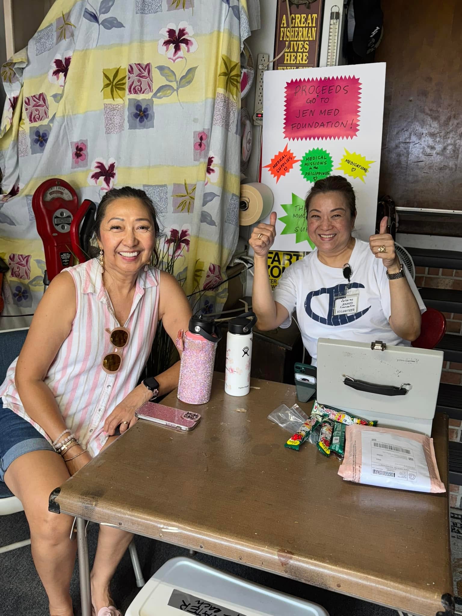 Two volunteers smile at a fundraising table, with a JenMed sign behind them.