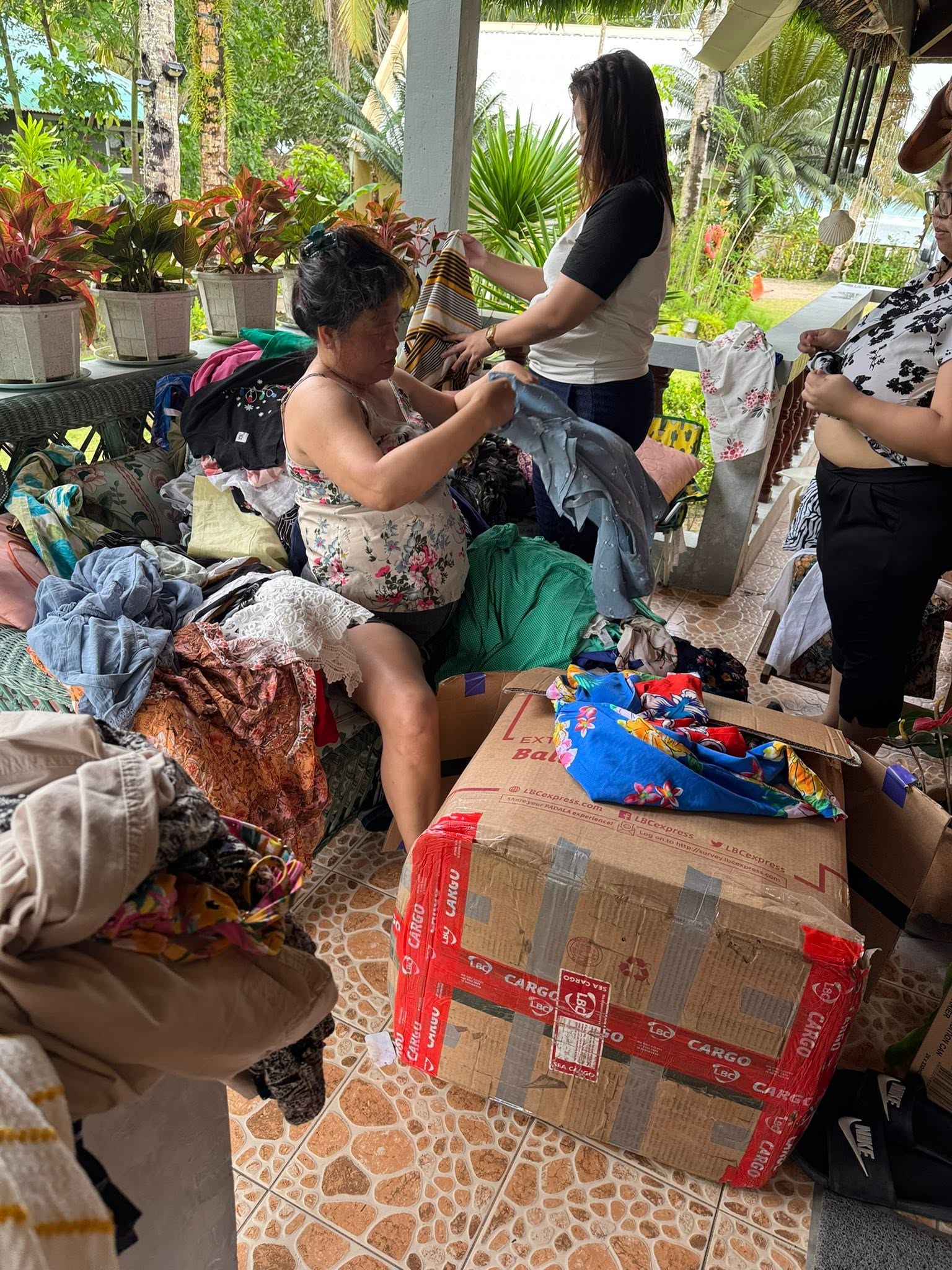 People sort clothing donations on a porch surrounded by plants.