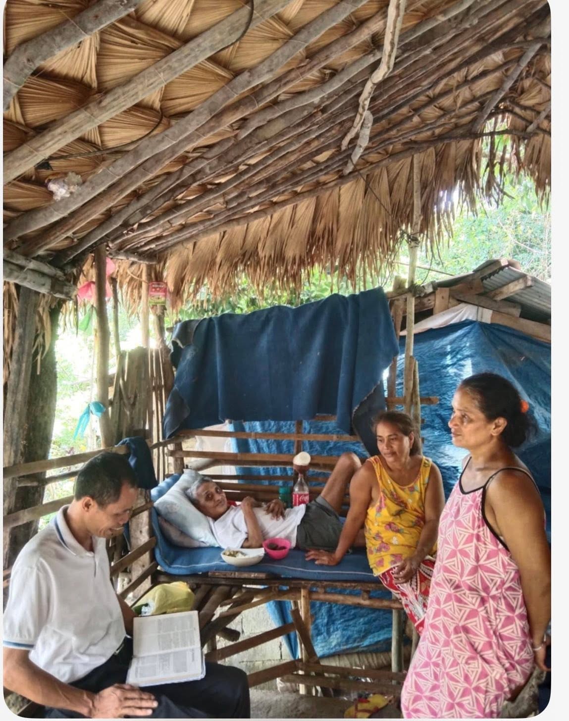 Visitors gather beside a person resting on a raised bed under a thatched roof.