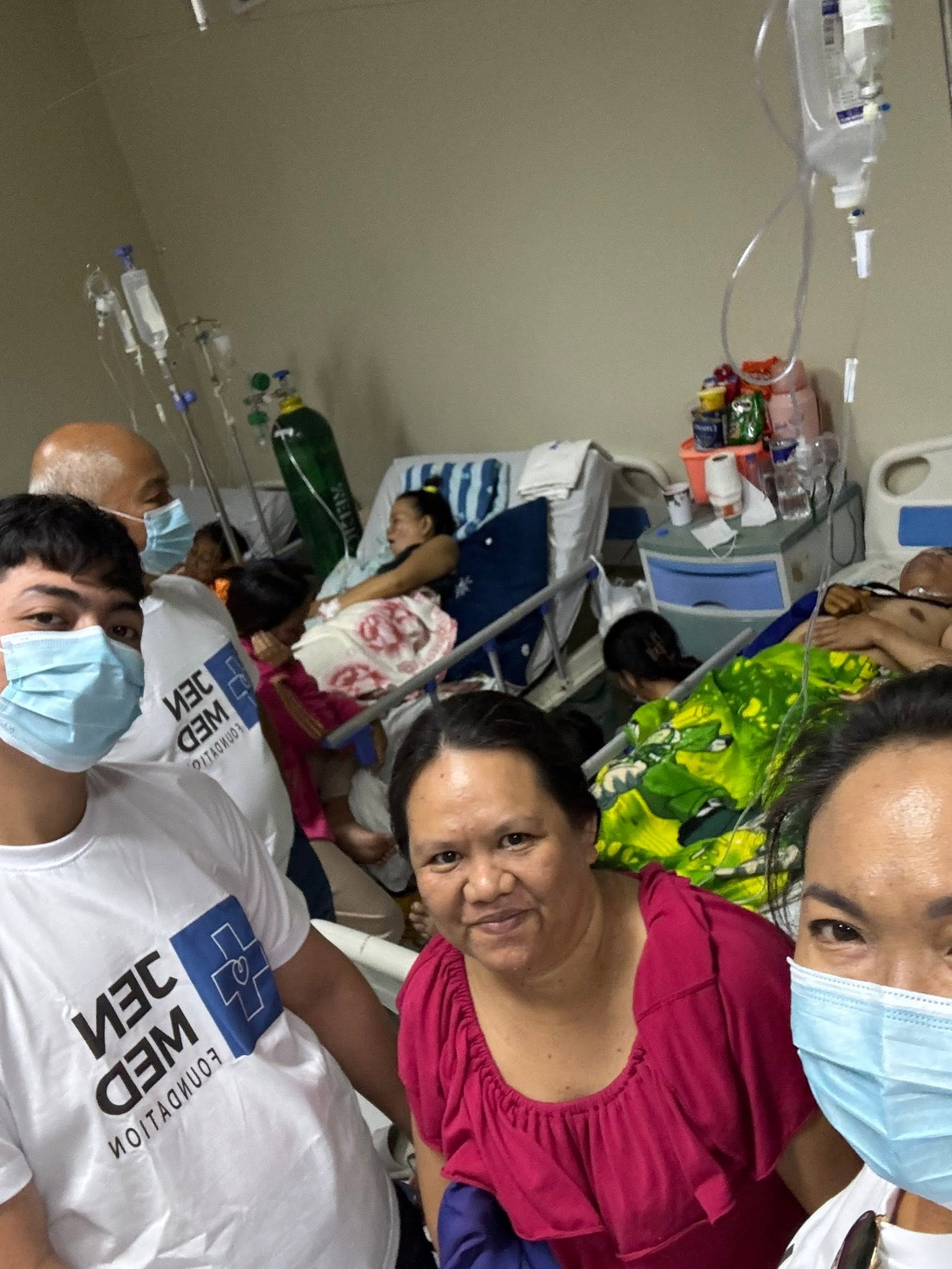 JenMed volunteers and visitors gather in a hospital room with patients resting behind them.
